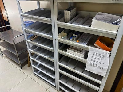 Estanterías metálicas en un área de almacenamiento médica, con cartel sobre entrega de medicamentos y un carrito metálico visible.
