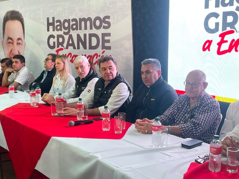 Arnoldo Wiens, con gestos tranquilos, se sienta rodeado de seis hombres y una mujer en mesa blanca y roja, atentos al evento político.