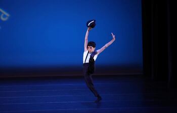 Joven bailarín en camiseta blanca y pantalones negros sostiene un sombrero mientras eleva una pierna, con fondo azul atractivo.