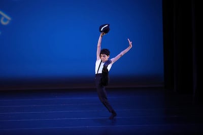 Joven bailarín en camiseta blanca y pantalones negros sostiene un sombrero mientras eleva una pierna, con fondo azul atractivo.