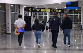 Cuatro personas en movimiento en un aeropuerto. Mujer con maleta, hombre de espaldas con objeto azul, ambiente luminoso.