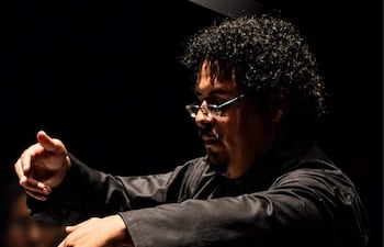 Luis Chumpitazi Montoya en chaqueta negra, dirigiendo con gestos en auditorio con músicos detrás.