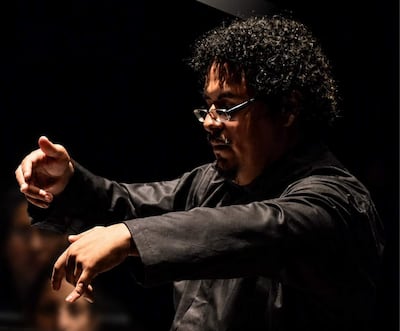Luis Chumpitazi Montoya en chaqueta negra, dirigiendo con gestos en auditorio con músicos detrás.