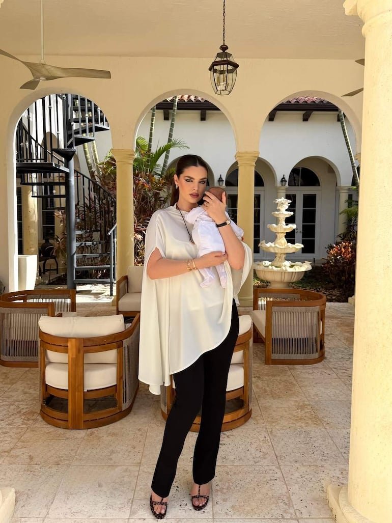 Nadia Ferreira con blusa blanca y pantalones negros sonríe sosteniendo a un bebé en brazos, en un patio iluminado.