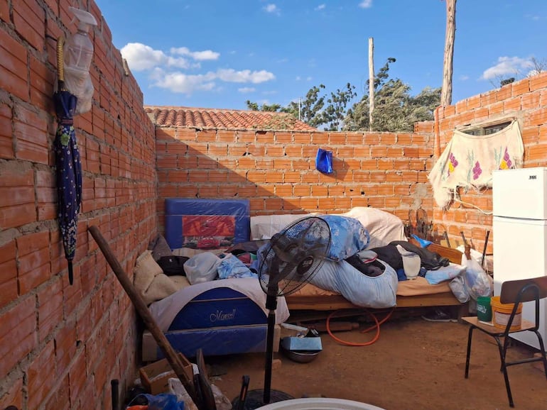 Interior de vivienda desordenada con paredes de ladrillo, sofá sin cojines y ropa en el suelo.