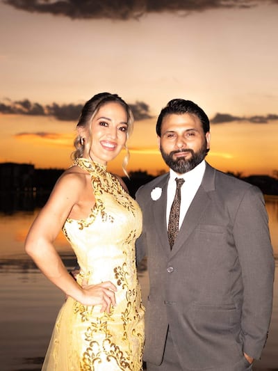 Mujer con vestido amarillo y hombre en traje gris sonríen frente a un atardecer sobre el agua, en un ambiente festivo.