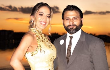 Mujer con vestido amarillo y hombre en traje gris sonríen frente a un atardecer sobre el agua, en un ambiente festivo.