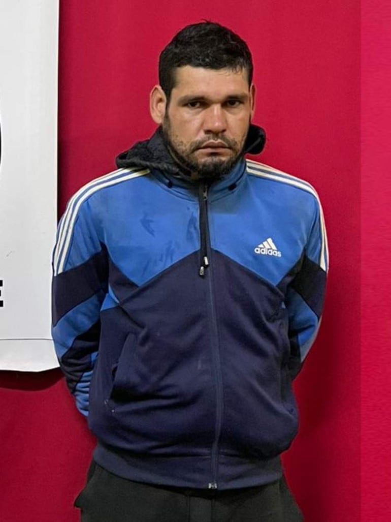 Hombre serio con chaqueta azul y franjas blancas, en fondo rojo, con cartel blanco parcialmente visible a la izquierda.