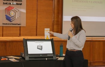 Mujer con camisa de rayas sostiene un micrófono mientras presenta documentos sobre elecciones en un auditorio con equipo técnico.