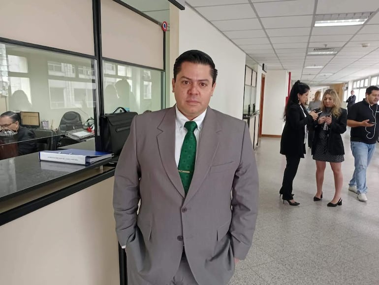 Hombre con traje gris y corbata verde de pie en el centro, rodeado de personas interaccionando en un ambiente administrativo.