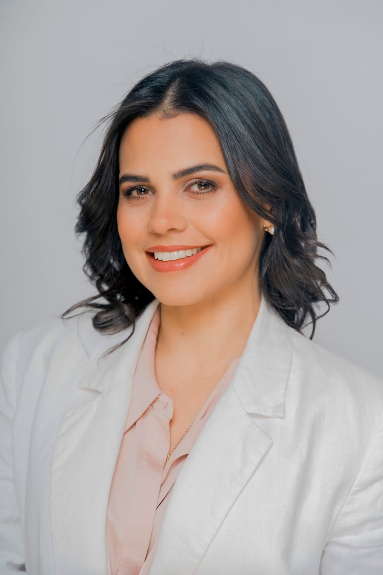 Dra. Bianca Parra Roig cabello oscuro y rizado, vestida con chaqueta blanca y blusa clara, sonríe en primer plano sobre fondo neutro.