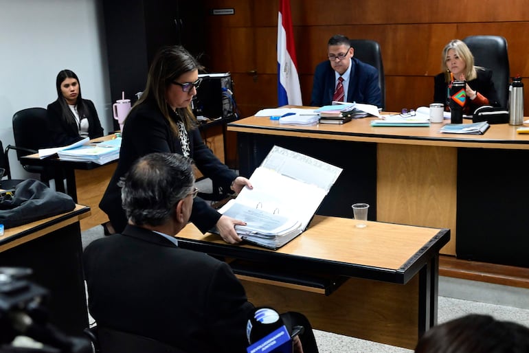 Mujer en el centro hablando y sosteniendo un expediente, rodeada de tres personas en un entorno formal con documentos y banderas.