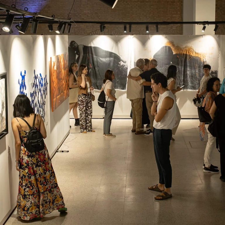 Asistentes de la feria se abrazan y observan obras de arte en un museo, rodeados de luces cálidas y coloridos atuendos.