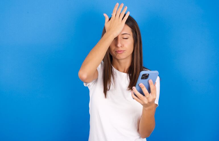 Una mujer joven se golpea la frente con una mano mientras sostiene en la otra un teléfono celular.