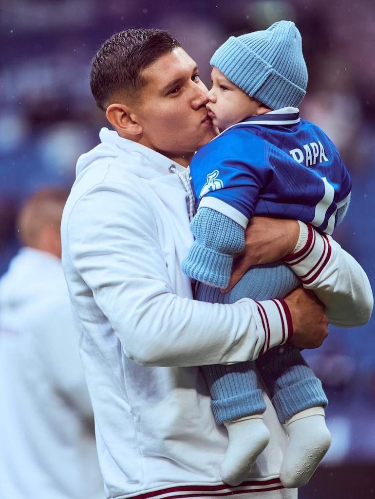 Juan Cáceres, con chaqueta blanca y rojo, besa a un niño con camiseta azul que dice 'PAPA' en un evento deportivo cálido.