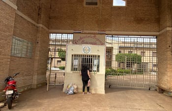 Mujer de espaldas con cabello largo y oscuro, sosteniendo un objeto, junto a una ventanilla de rejas en un ambiente penitenciario.
