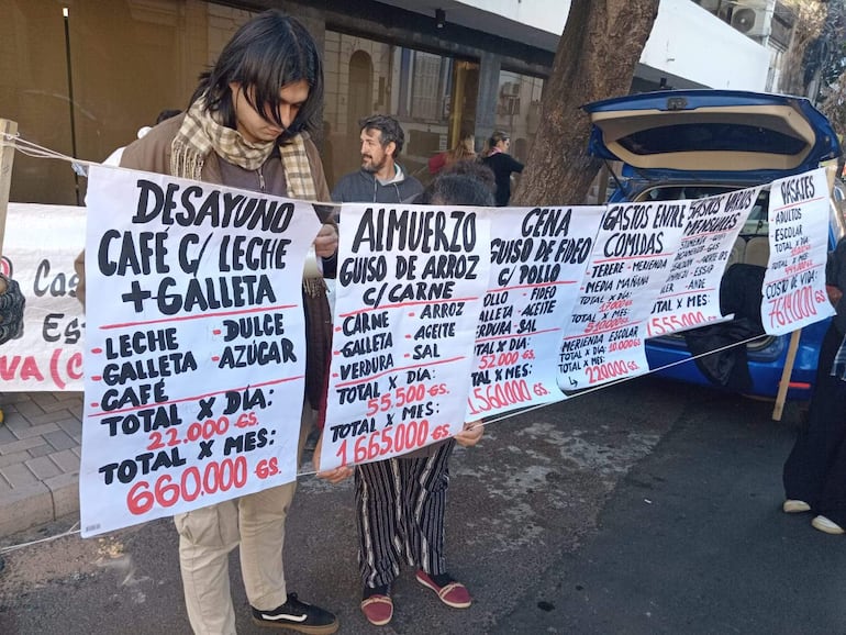 Personas sosteniendo carteles con menú y precios de comidas, en un ambiente exterior durante la manifestación.