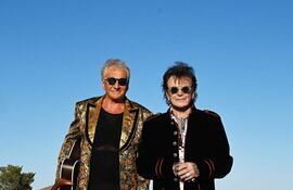 Dos hombres del dúo Air Supply, uno con guitarra brillante y otro en abrigo negro, posan en una carretera desértica con montañas al fondo.