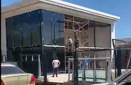 Trabajadores en andamio y en el suelo, vestidos con ropa de trabajo, en un edificio con paneles de vidrio en construcción.