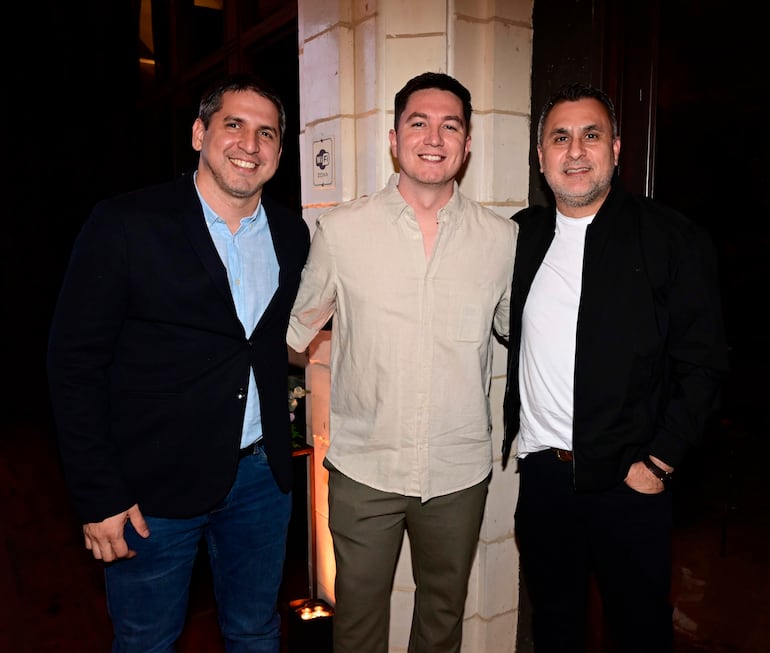 Tres hombres sonrientes: uno en blazer negro y camisa azul, otro con camisa clara y el tercero en camiseta blanca bajo chaqueta negra.