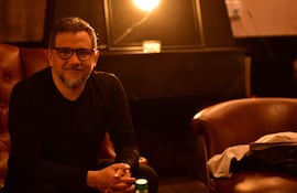 Músico de cabello gris y lentes sentado en un sillón marrón, con iluminación tenue y botella de agua en la mesa.