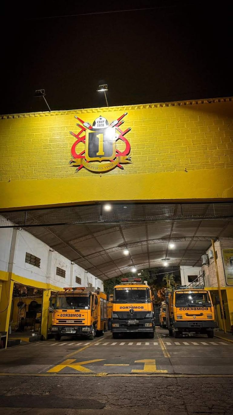 Cuartel de Bomberos K1 con cuatro camiones amarillos alineados, iluminados y emblema de la compañía visible.