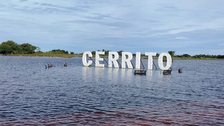 Letras grandes que dicen 'CERRITO', parcialmente sumergidas en agua, rodeadas de vegetación y cielo despejado.