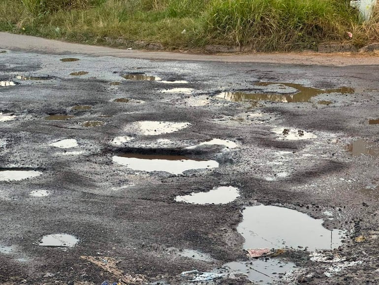 Calle deteriorada con profundos baches y charcos de agua acumulada, reflejando la luz del sol.
