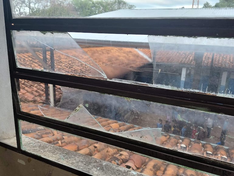 Marco de ventana roto con fragmentos de vidrio. Personas observan el daño afuera. Ambiente rural con techos de cerámica.