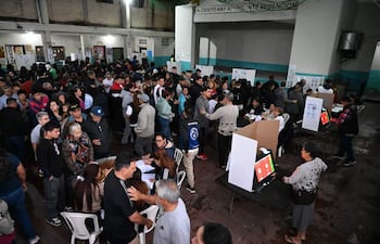 Recinto electoral lleno de personas organizadas en filas, con mesas de votación y pantallas visibles con opciones de votos.