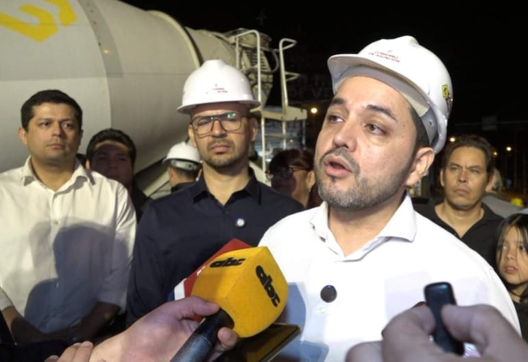 Luis Bello con micrófono en mano, rodeado de hombres en cascos de seguridad, discutiendo sobre la reparación de calles en un ambiente de trabajo.