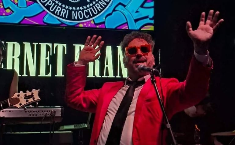 Carlos Colombino en traje rojo y gafas naranjas, sosteniendo un micrófono y saludando al público desde el escenario.