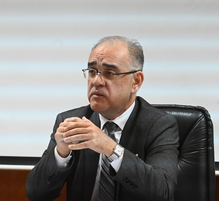 Humberto Otazú, con cabello canoso y gafas, gesticula mientras habla en un entorno formal, vistiendo chaqueta oscura y corbata a rayas.