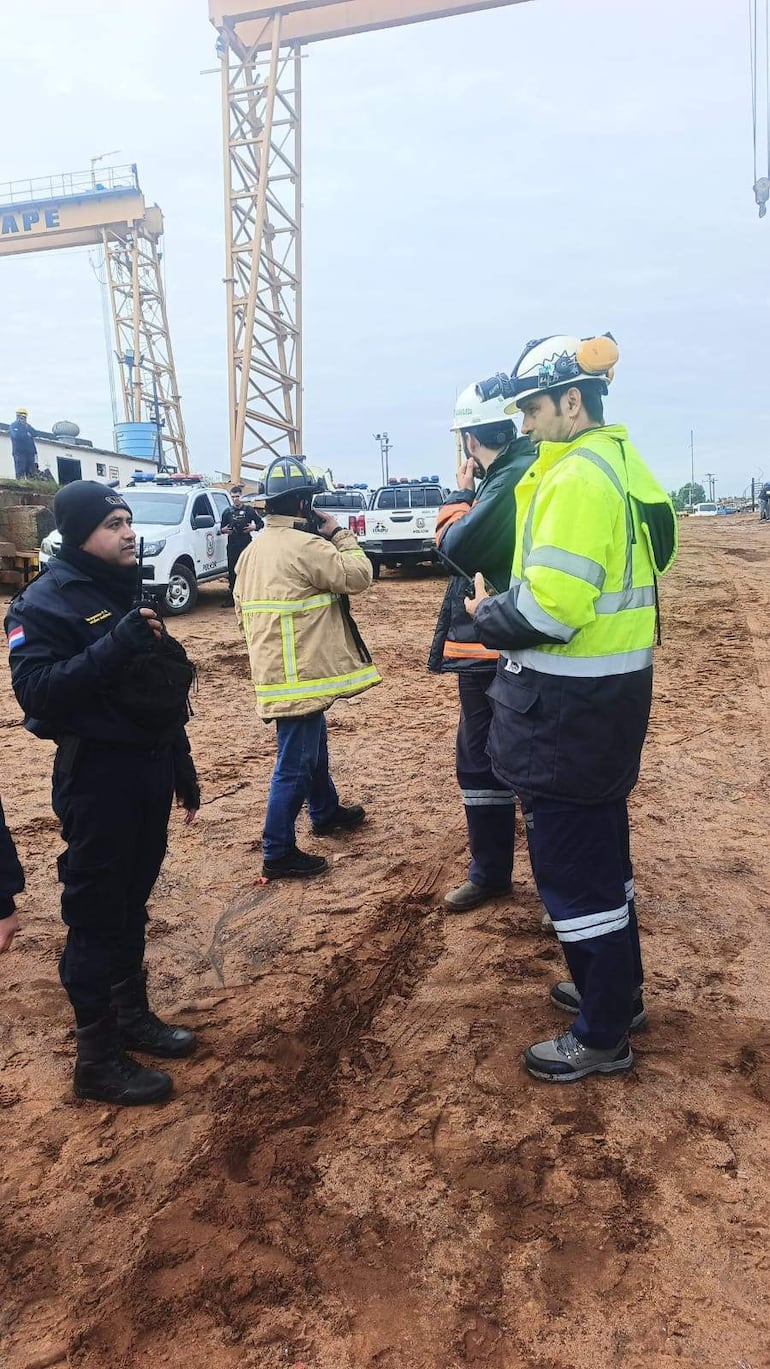 Cinco personas en vestimenta de seguridad, dos conversan mientras un hombre con casco negro observa en un entorno industrial bajo cielo nublado.