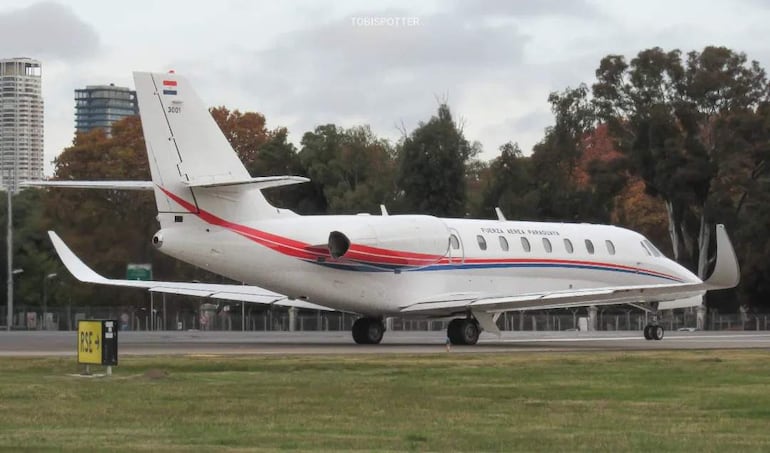 Avión blanco con bandas rojas y azules en pista de aterrizaje, rodeado de vegetación y edificios.