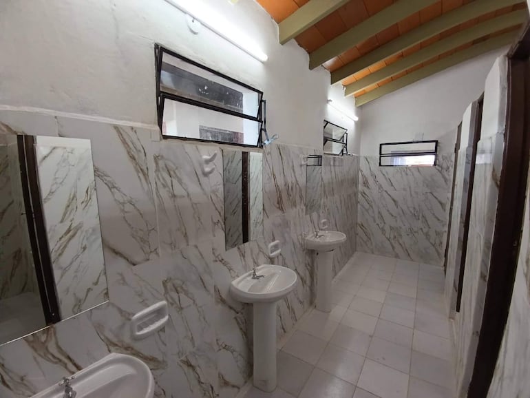 Baño escolar con tres lavamanos de pedestal y azulejos en tonos beige, iluminado por tubo fluorescente.