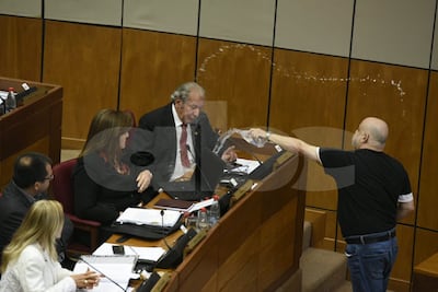 Hombre de camiseta negra lanza agua mientras el presidente de la sesión, con expresión de sorpresa, lo observa.