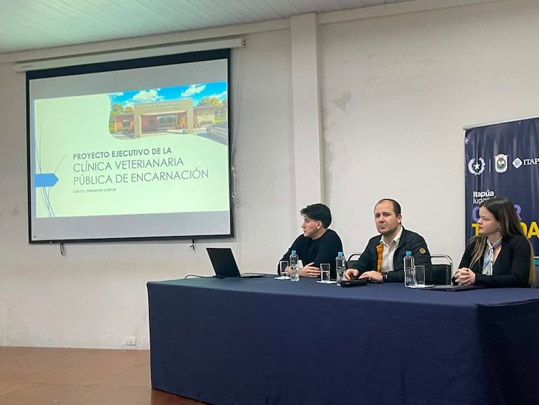 Tres personas en mesa de presentación; hombre central discutiendo proyecto, mientras otros dos escuchan atentamente en ambiente formal.