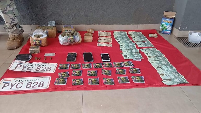 Objetos sobre manta roja, incluyendo paquetes de marihuana, billetes y teléfonos móviles, con parte de un militar al fondo.
