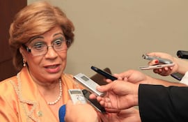 Gladys Bareiro de Módica con blusa naranja y accesorios, rodeada de periodistas con grabadoras en un ambiente interior.