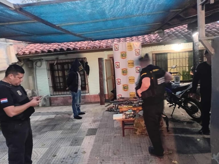 Hombre con chaleco táctico revisa su teléfono en operativo policial, debajo de un toldo azul y cerca de una moto.