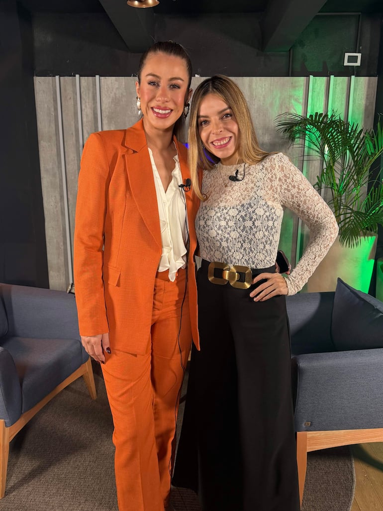 Jéssica Torres en traje naranja y Belén Bogado en blusa de encaje, de pie en un set moderno, sonrientes con micrófonos.