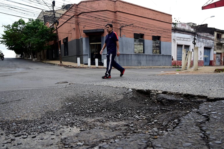 Hombre con camiseta oscura y pantalones deportivos camina por una calle con bache, rodeado de edificios y árboles.