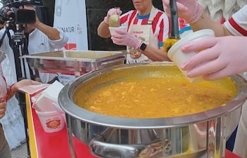 Mujer con guantes de látex y camiseta blanca sirve vori vori en recipiente de cartón, rodeada de personas y cámaras.