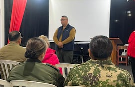 Hombre con chaleco marrón habla ante cinco personas sentadas, una mujer con abrigo rosa y otra con uniforme militar en un espacio decorado en rojo y negro.
