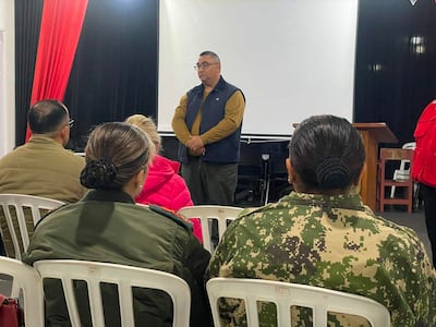 Hombre con chaleco marrón habla ante cinco personas sentadas, una mujer con abrigo rosa y otra con uniforme militar en un espacio decorado en rojo y negro.
