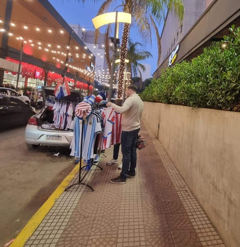 Hombre en suéter claro junto a carro blanco con camisetas de fútbol, mientras otro observa. Árboles y luces decorativas en el fondo.