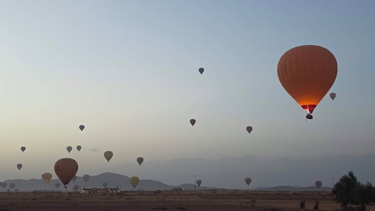 Globos aerostáticos de diversos colores en el cielo, destacando uno naranja con rojo, sobre un área de despegue iluminada.