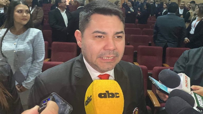 Hombre de traje oscuro y corbata roja habla a la prensa, observando una mujer con blusa gris, en un ambiente de auditorio.