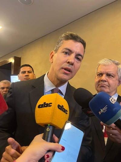 Santiago Peña en traje negro habla al micrófono, mientras Enrique Riera lo escucha atentamente en un ambiente formal.